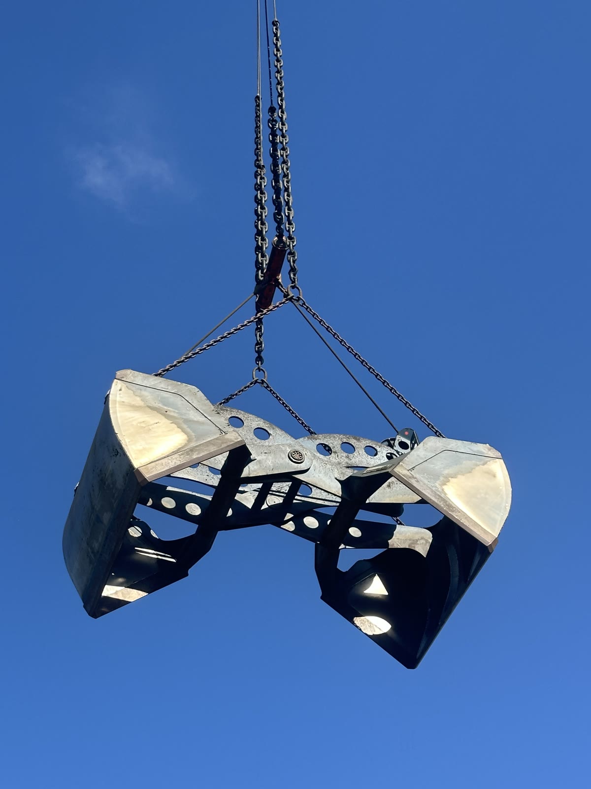 Cuchara bivalva portuaria completa suspendida de las cadenas de la grúa, contra el cielo despejado