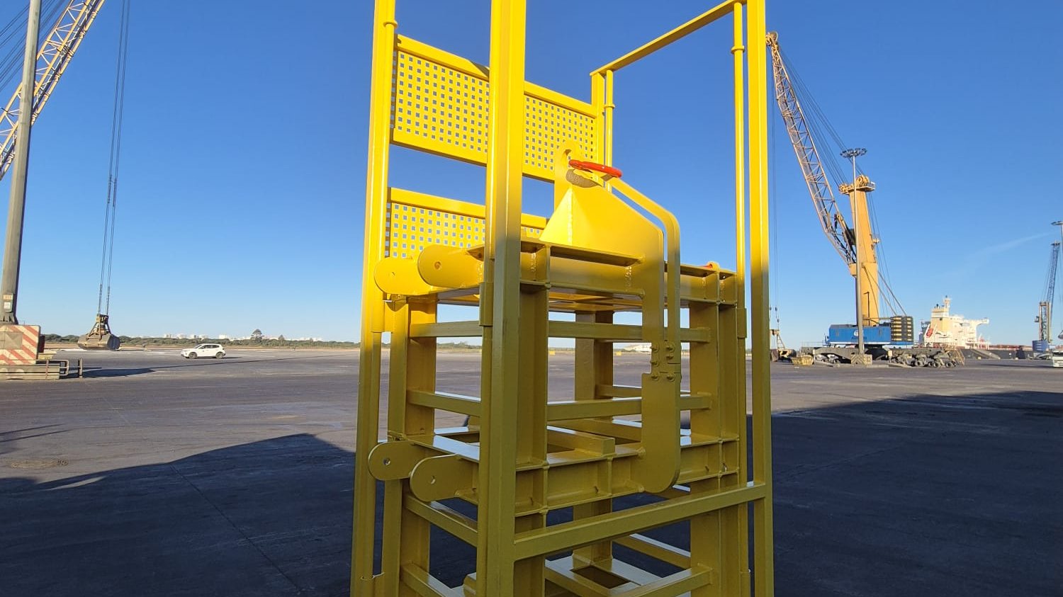 Cesta de trabajo recién fabricada y pintada en el muelle, con grúas portuarias y un buque al fondo