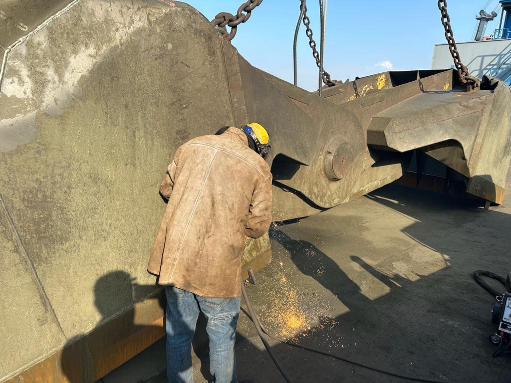 Reparación in situ de una cuchara bivalva portuaria a pie de muelle, con chispas de soldadura saltando